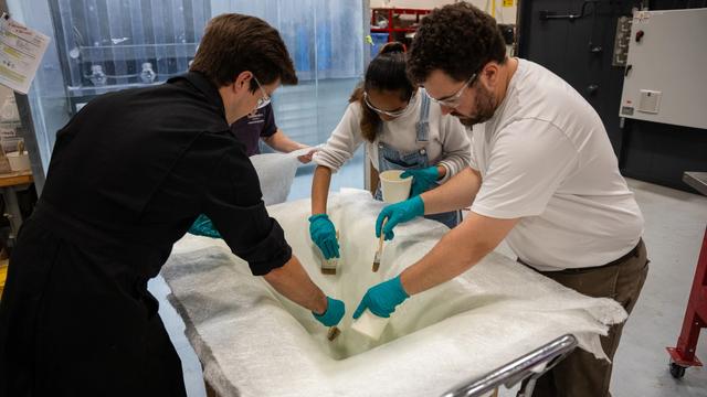 Three people wearing safety glasses and gloves brush resin onto a large white fabric piece in a workshop