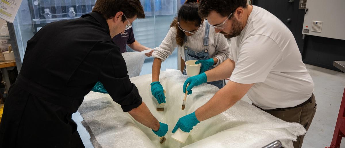 Three people wearing safety glasses and gloves brush resin onto a large white fabric piece in a workshop