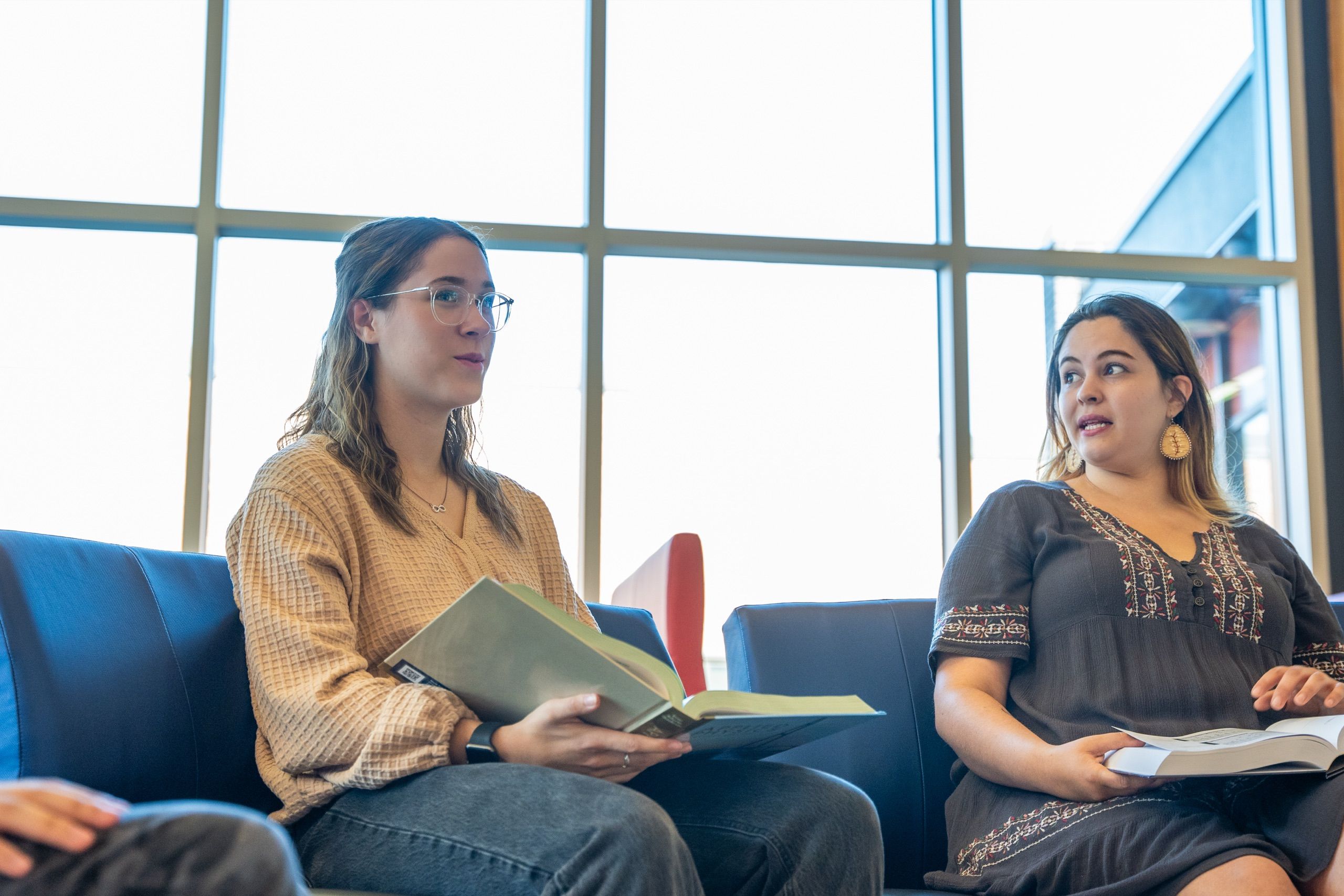 Two seated women hold open books in a bright library lounge with blue chairs and large windows.