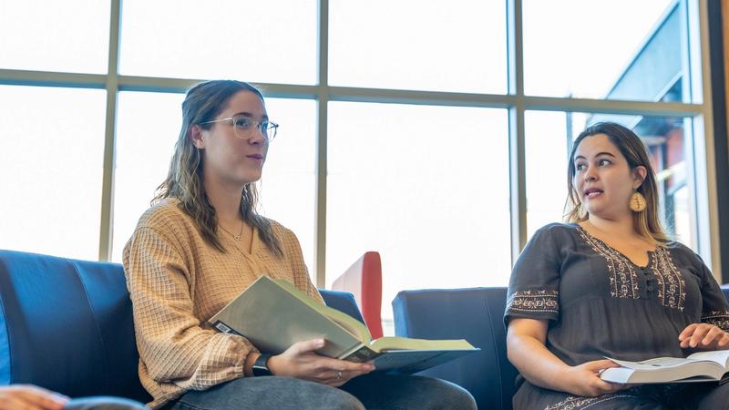 Two seated women hold open books in a bright library lounge with blue chairs and large windows.