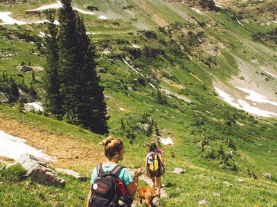 People hiking on a mountain trail through alpine terrain with patches of snow and evergreen trees