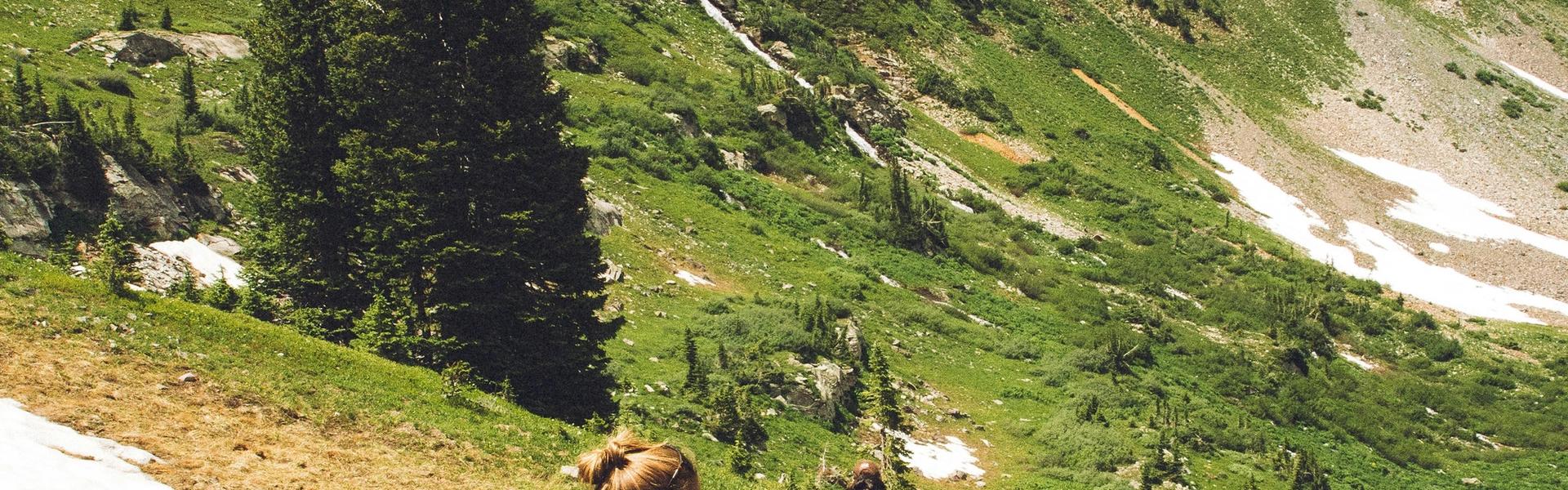 People hiking on a mountain trail through alpine terrain with patches of snow and evergreen trees