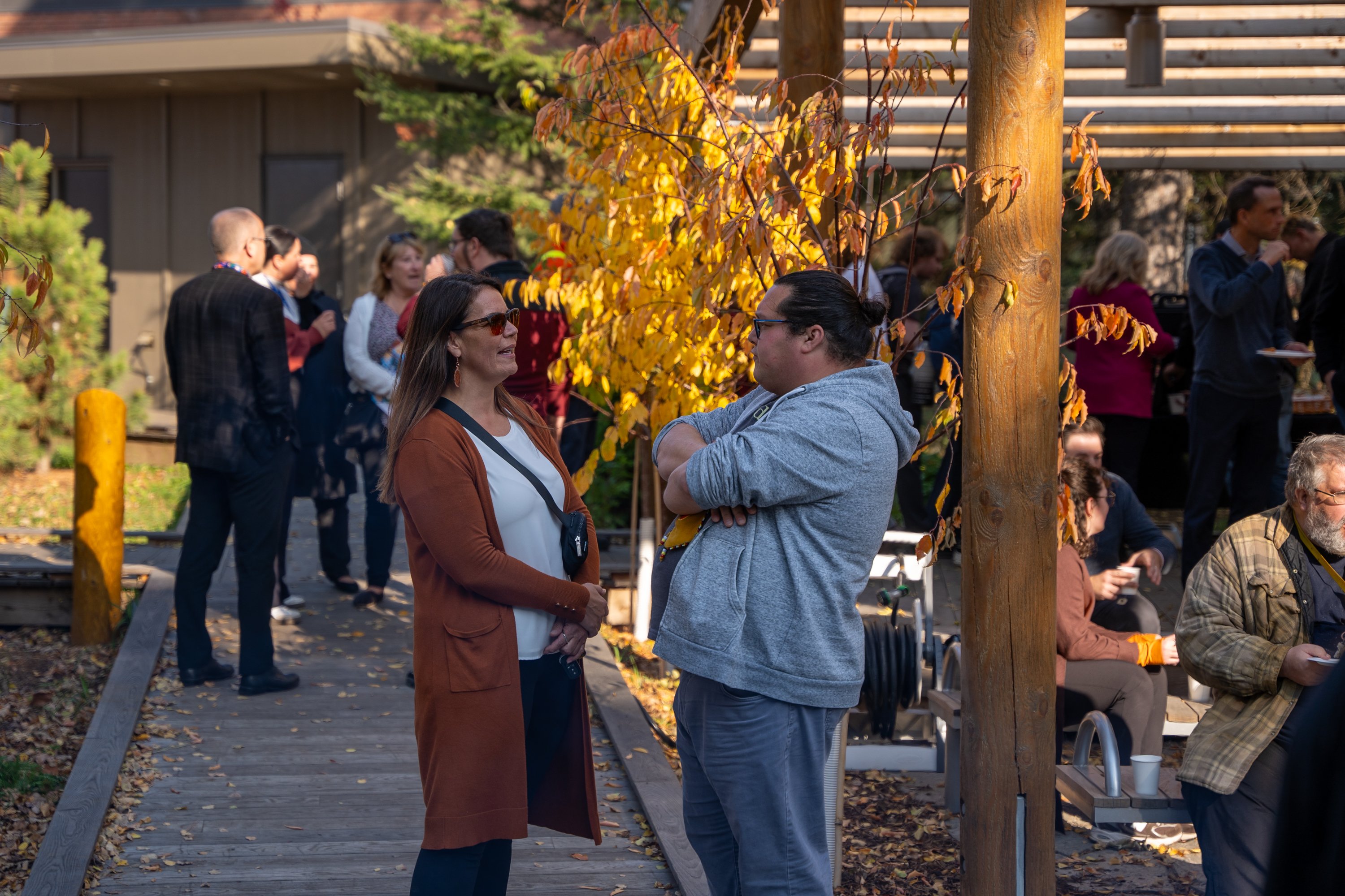 Two people conversing at an outdoor campus gathering, with others socializing and autumn foliage in the background.