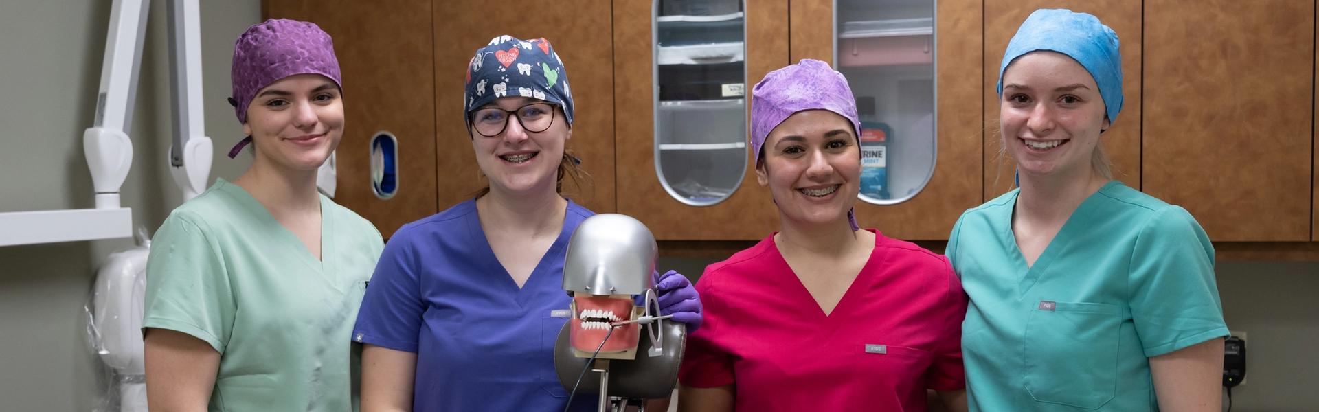 Four dental students in scrubs and colorful caps standing around a dental chair in a clinic setting at Confederation College.