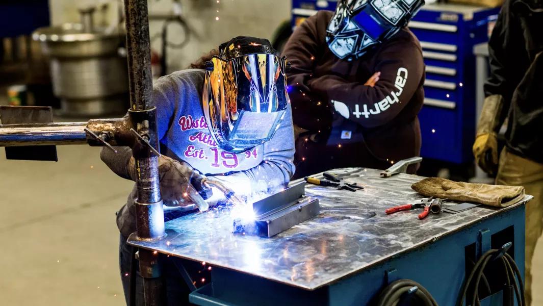 Welding students practicing metal fabrication at a workbench using protective helmets