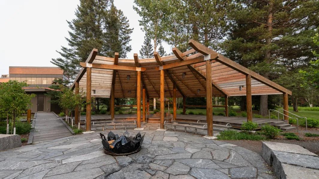 Wooden pavilion with fire pit and seating surrounded by trees on campus