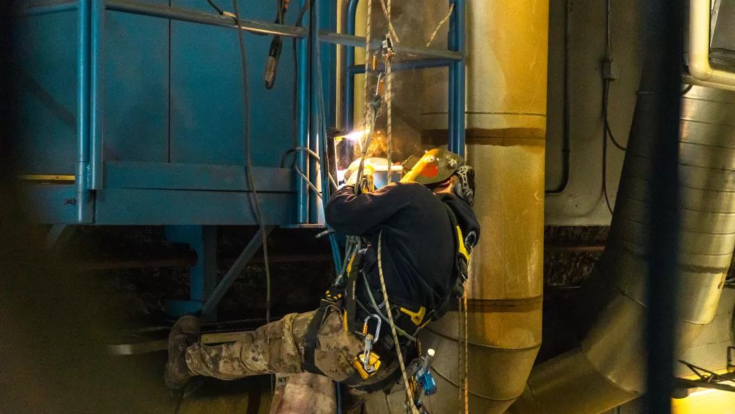 Industrial worker welding while suspended in safety gear inside a large facility