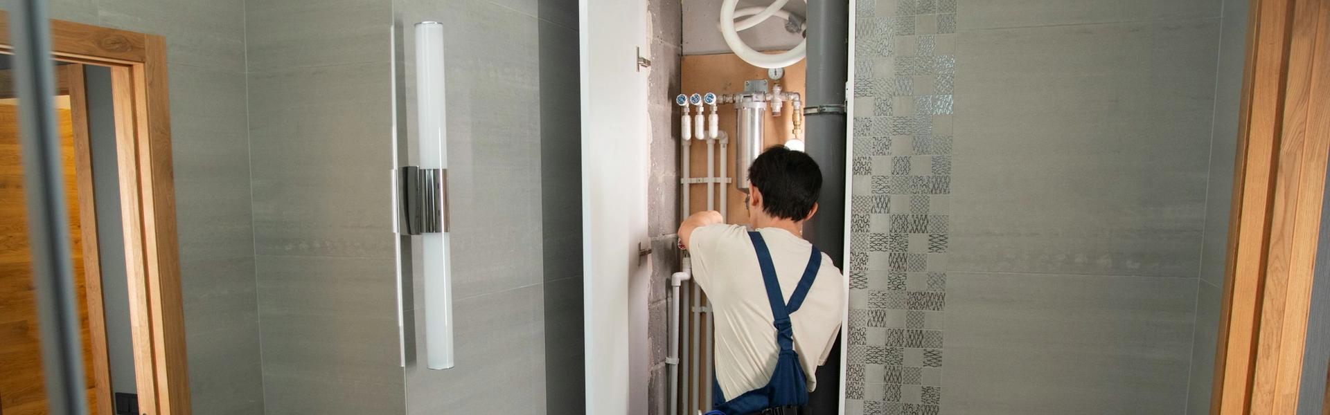 Plumber repairing exposed pipes behind wall panel above toilet in a bathroom.