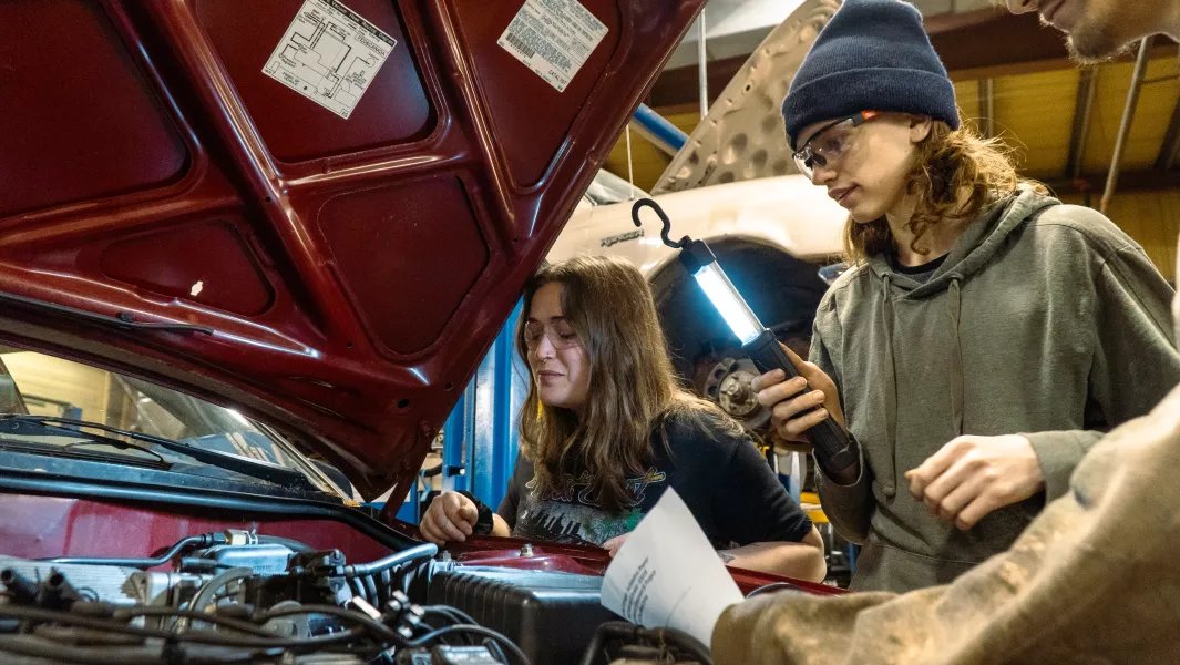 Students examining an engine bay together under the raised hood of a vehicle