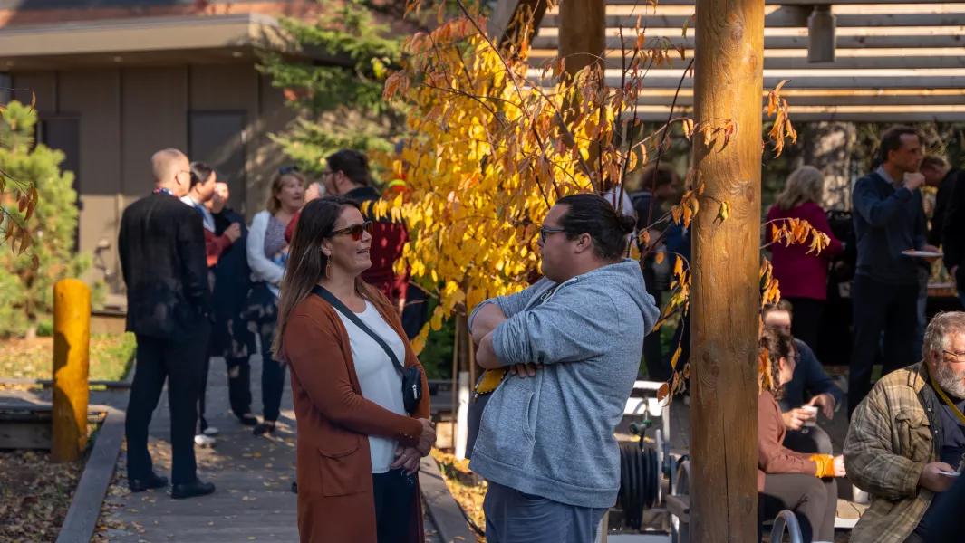 Two people chatting outdoors at a fall gathering, with yellow autumn leaves and a small crowd in the background