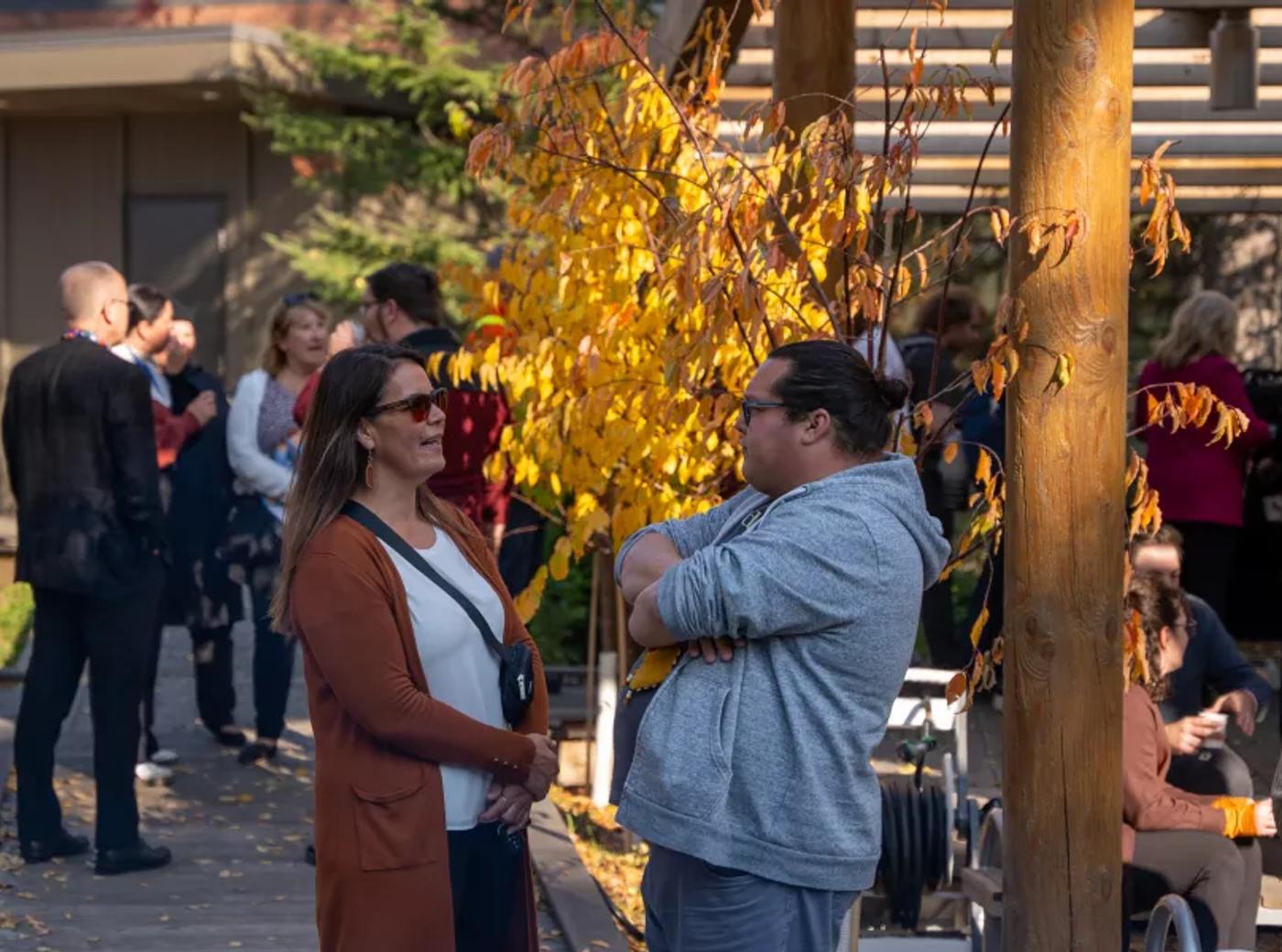 Two people chatting outdoors at a fall gathering, with yellow autumn leaves and a small crowd in the background