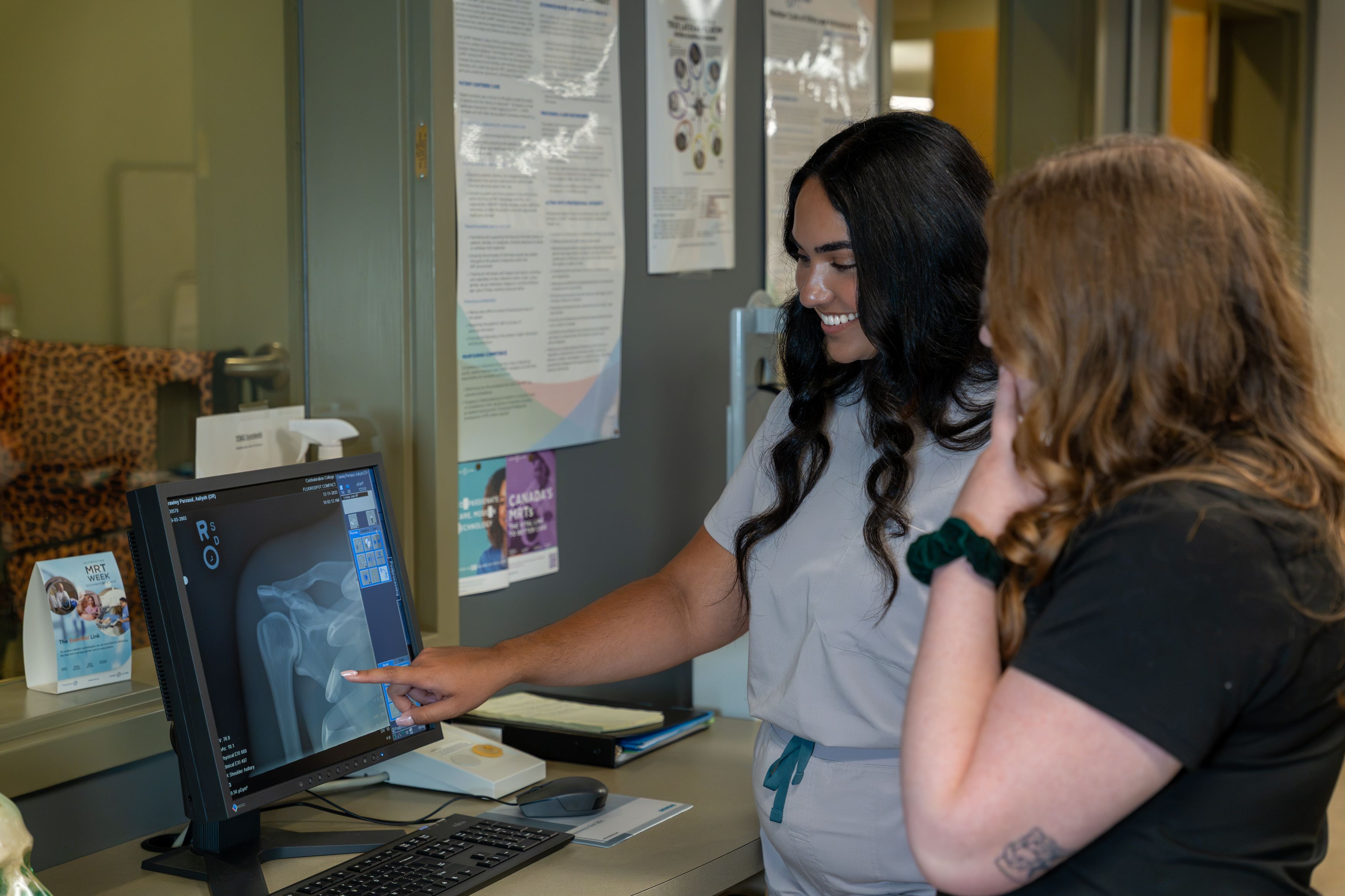 Two people examine a pelvic X-ray on a computer monitor; one points to the image in a medical imaging room.