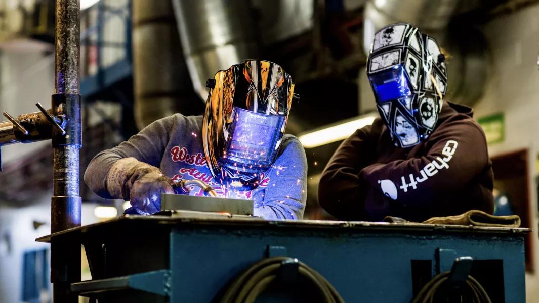 Student welding metal at a bench while a classmate observes nearby
