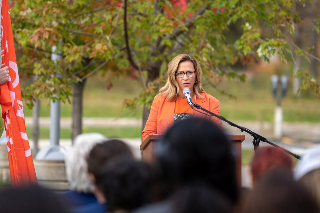 A woman in an orange jacket speaks into a microphone at a wooden podium outdoors, with an audience and trees behind her.