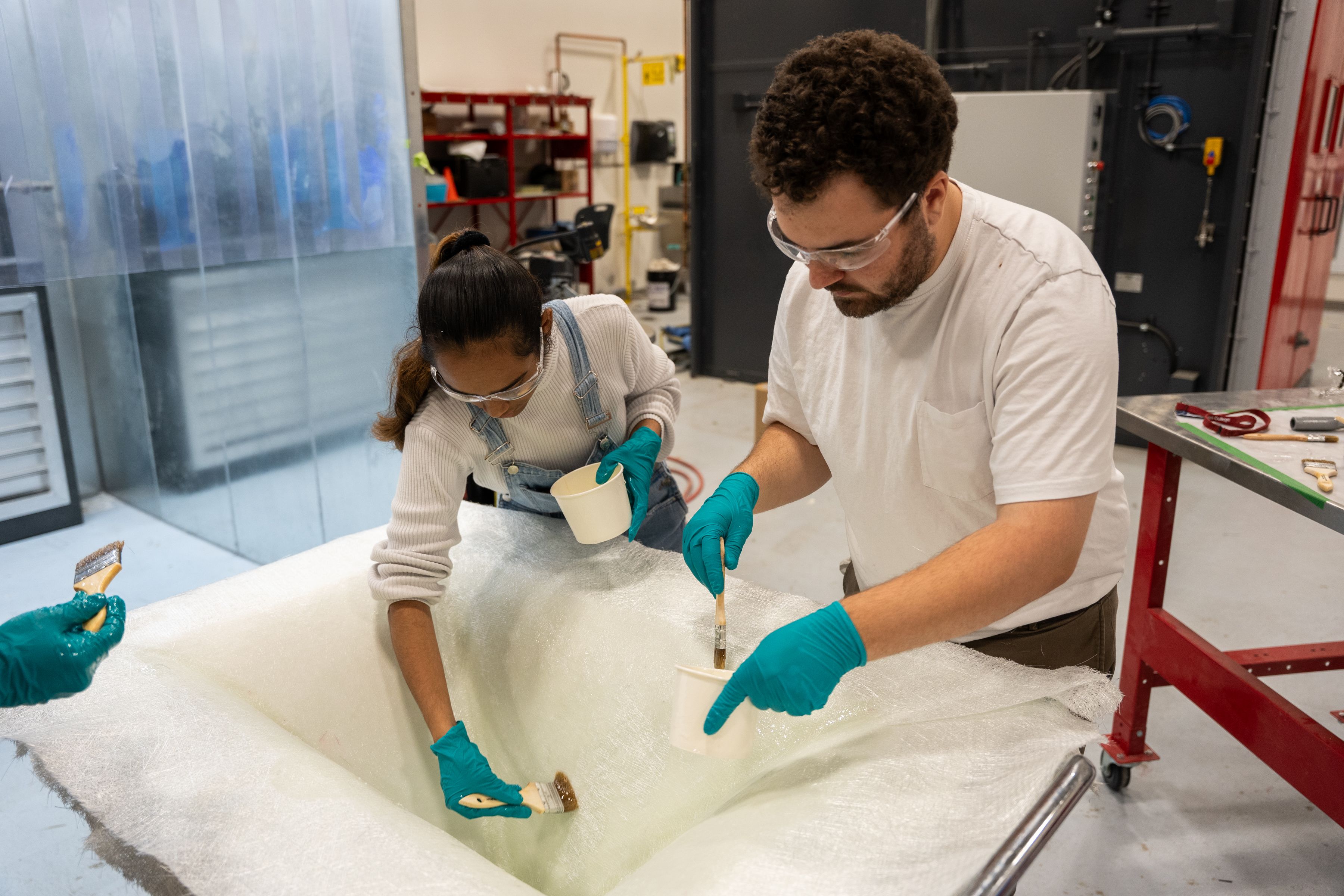 Two people wearing safety glasses and blue gloves brush a white coating onto a large foam surface in a workshop