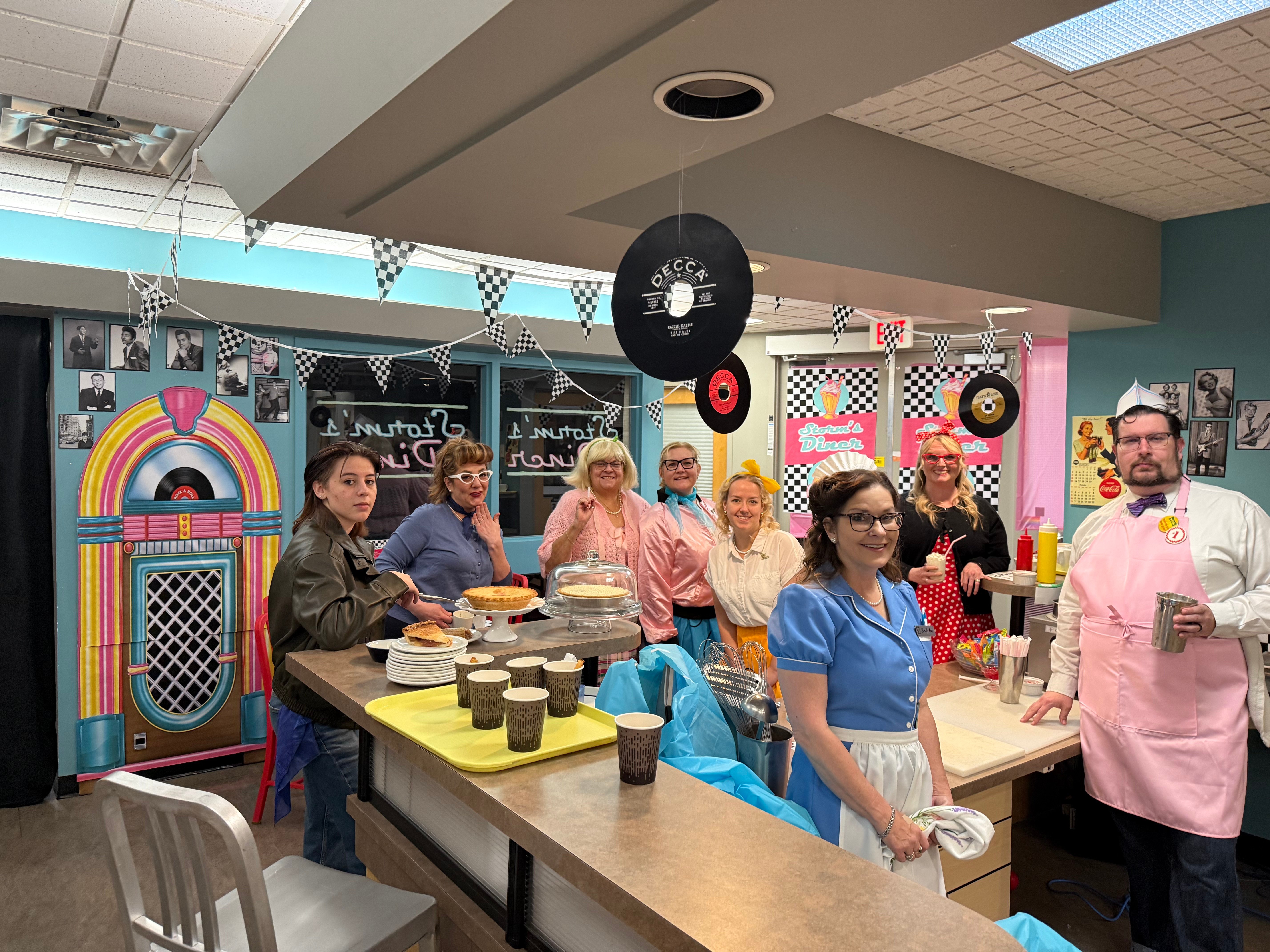 People in colourful costumes pose behind a counter of snacks in a diner decorated with records and checkered flags.