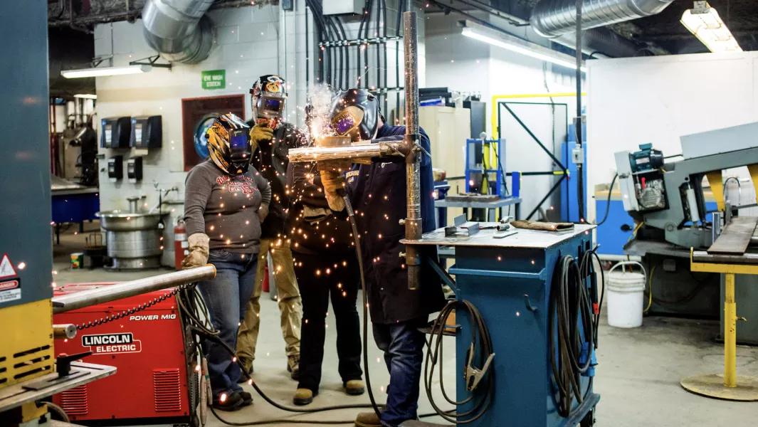 Welding students collaborating on a metal joint while sparks fly in a lab setting
