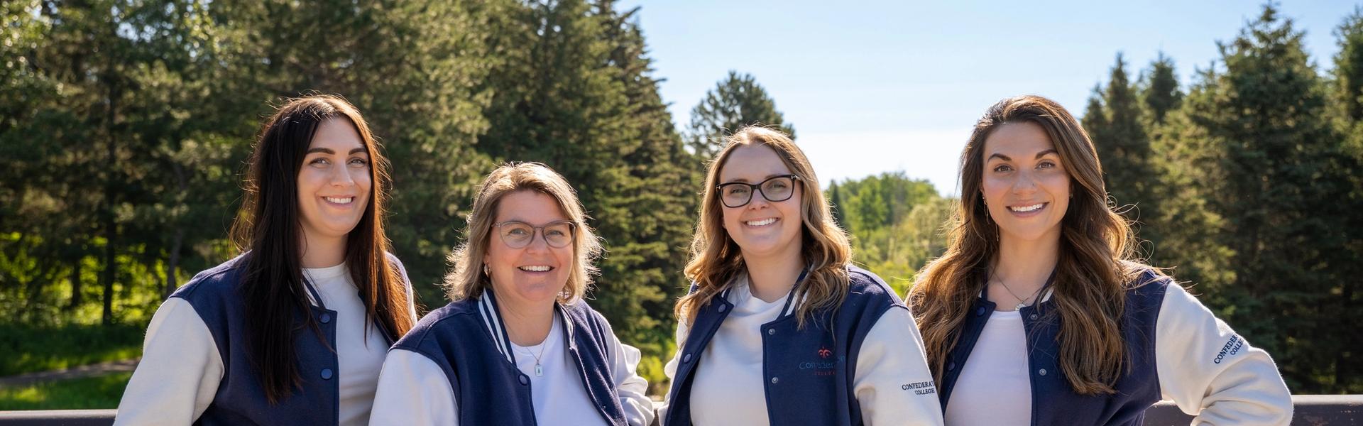 Four women standing outdoors in matching navy-and-white varsity jackets