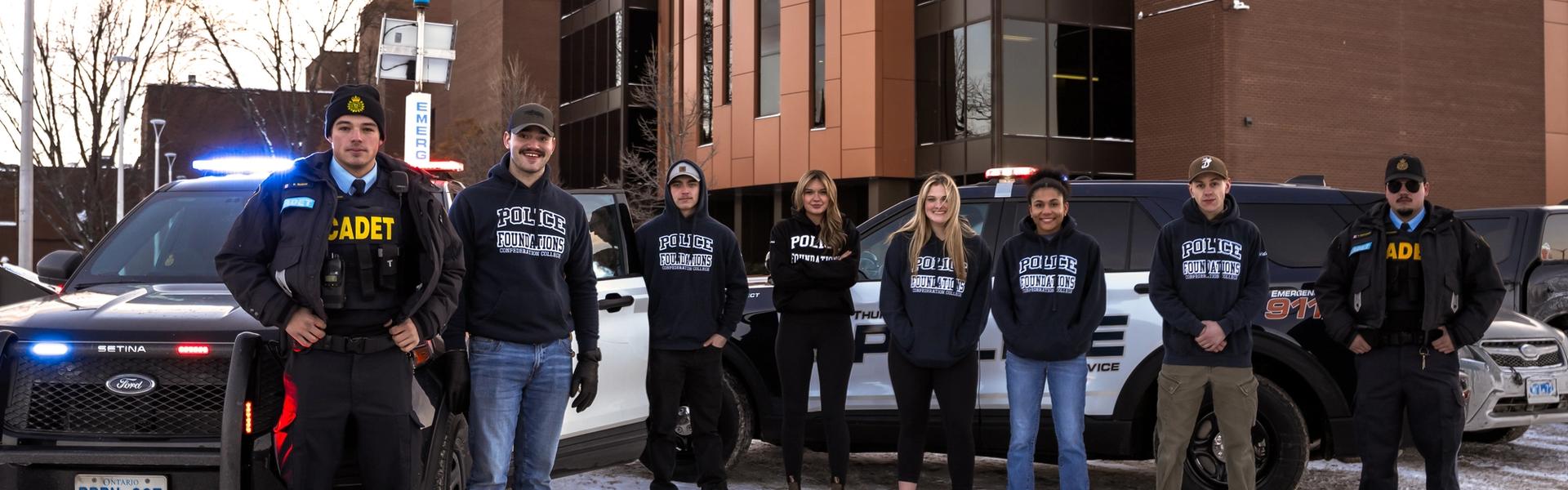 Police foundations students and cadets standing with patrol vehicles outside a campus building in winter.