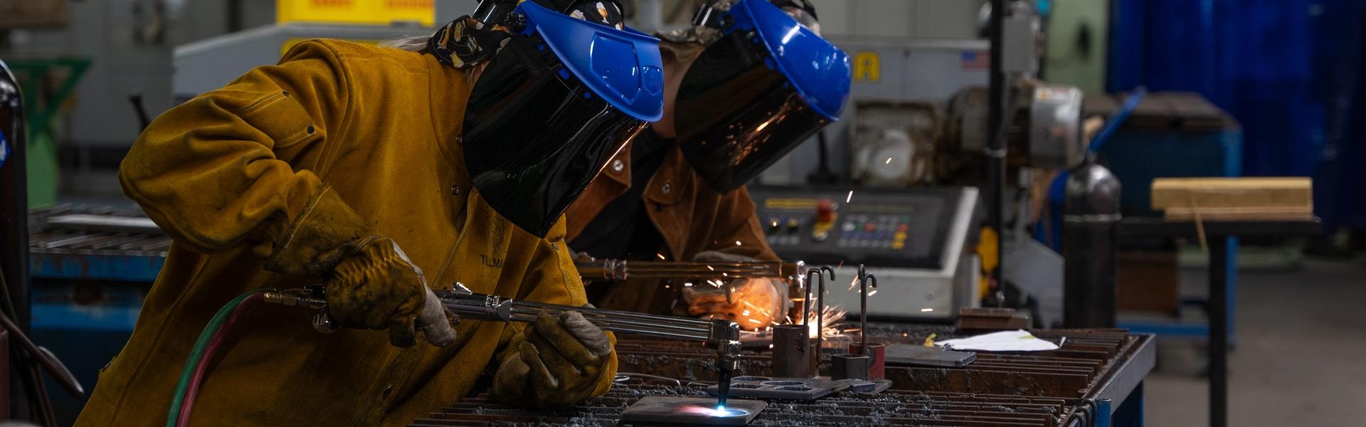 Two students wearing welding helmets and heavy protective jackets work at a metal welding station in a trades lab