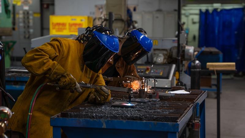 Two students wearing welding helmets and heavy protective jackets work at a metal welding station in a trades lab