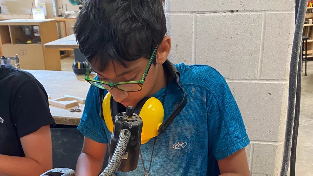 Young student using a drill press in a woodworking workshop during summer camp