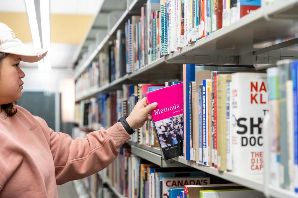 A student in a white cap and pink sweatshirt reaches for a bright pink textbook on a library shelf. Rows of books fill the aisle in a quiet campus library setting