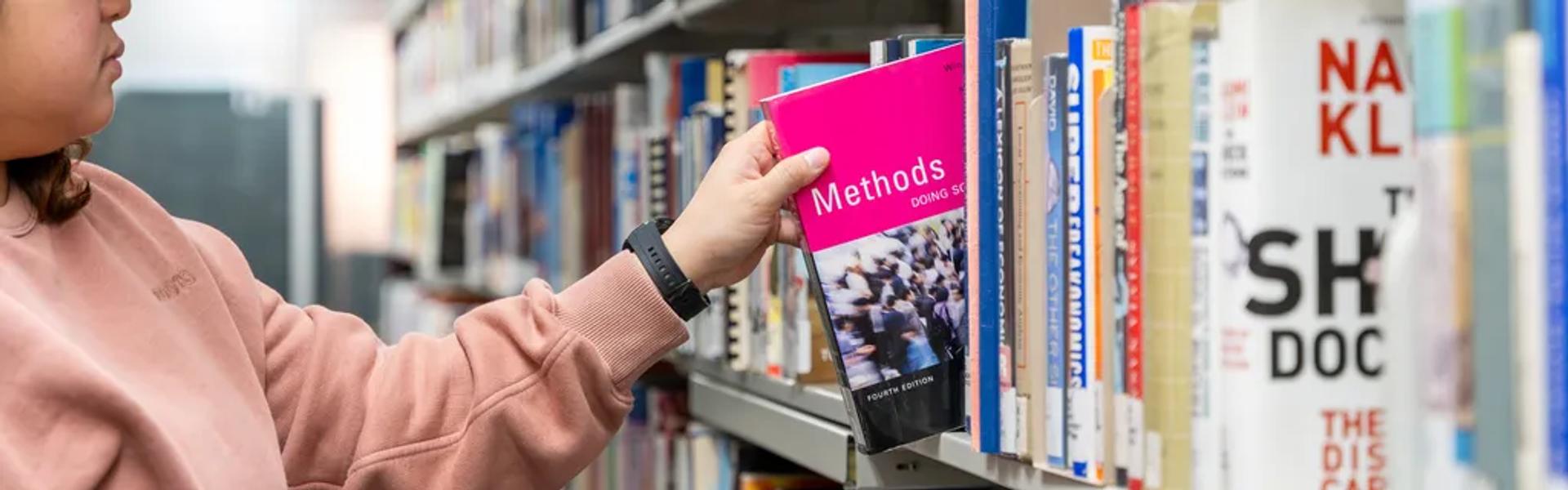 A student in a white cap and pink sweatshirt reaches for a bright pink textbook on a library shelf. Rows of books fill the aisle in a quiet campus library setting
