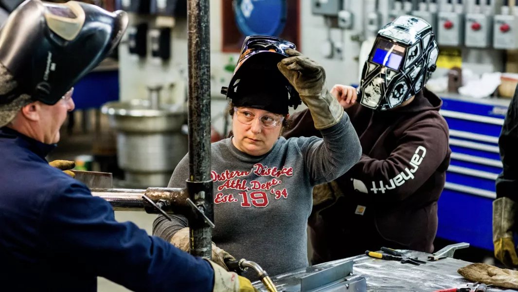 Welding students adjusting safety helmets and reviewing work with an instructor