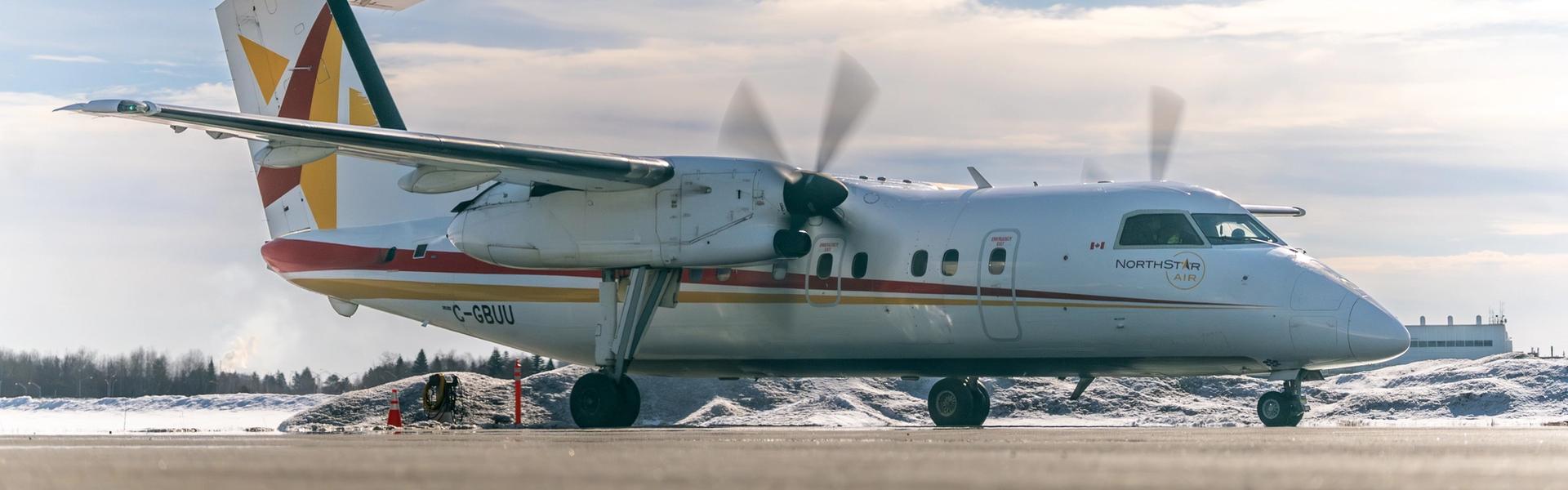 A white North Star Air turboprop airplane sits on a snowy runway under a bright sky. The aircraft is shown in profile with its propellers blurred from motion and colored stripes on the tail and fuselage