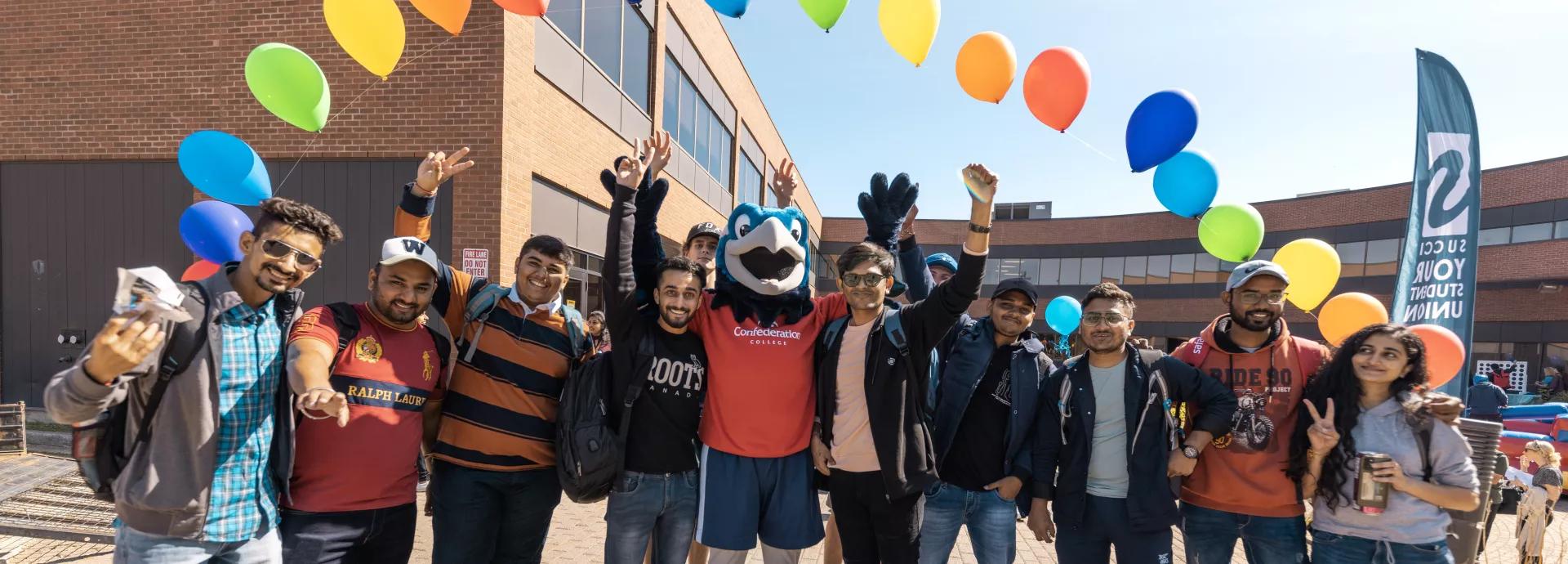 Students celebrate with the Confederation College mascot on campus under colorful balloons