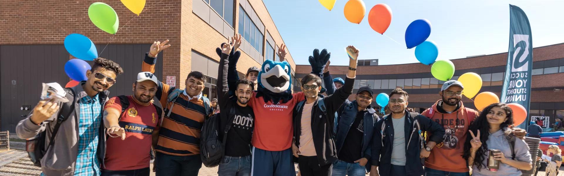 Students celebrate with the Confederation College mascot on campus under colorful balloons