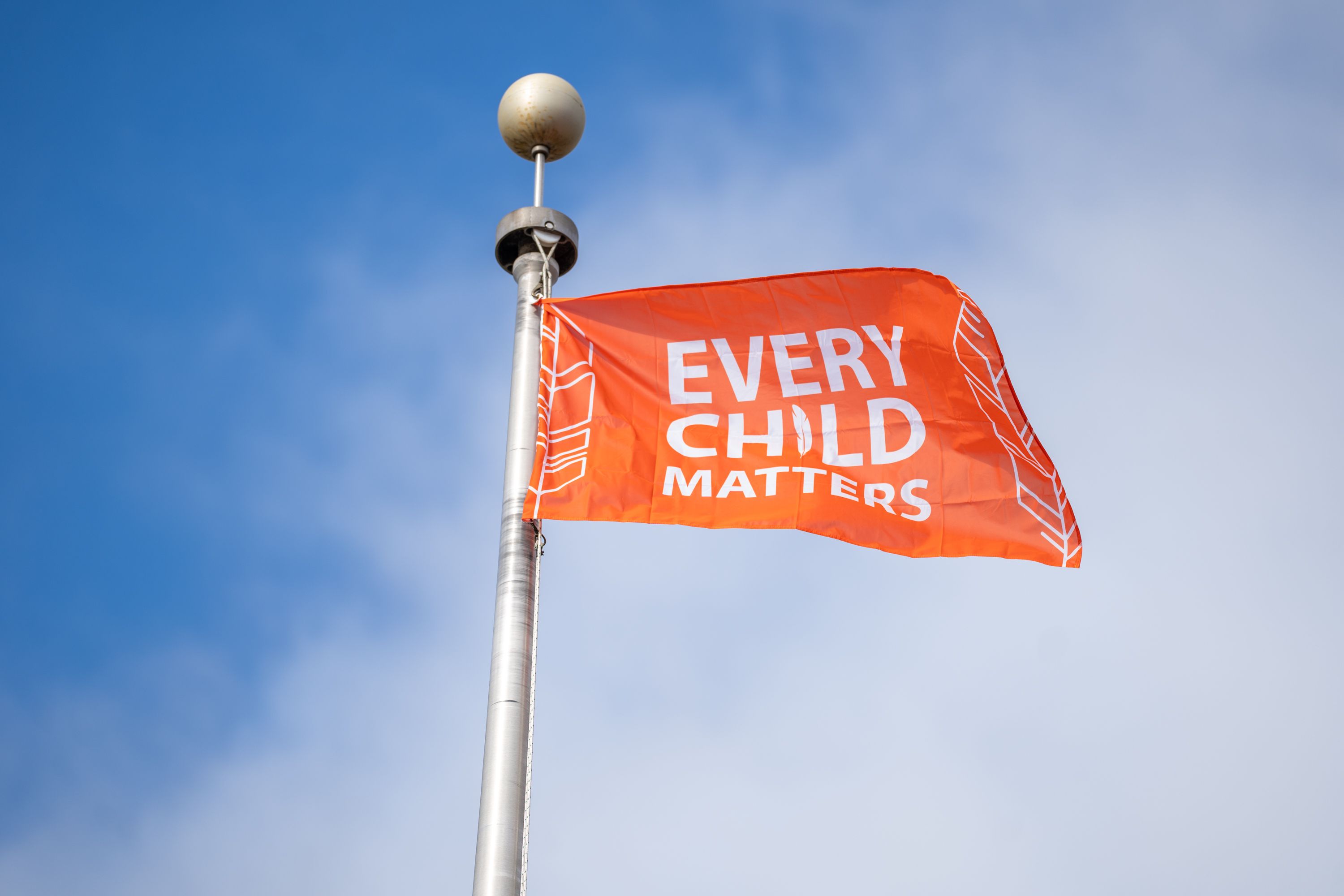 An orange flag reading “Every Child Matters” flies from a metal pole against a blue sky.
