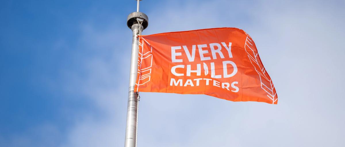 An orange flag reading “Every Child Matters” flies from a metal pole against a blue sky.
