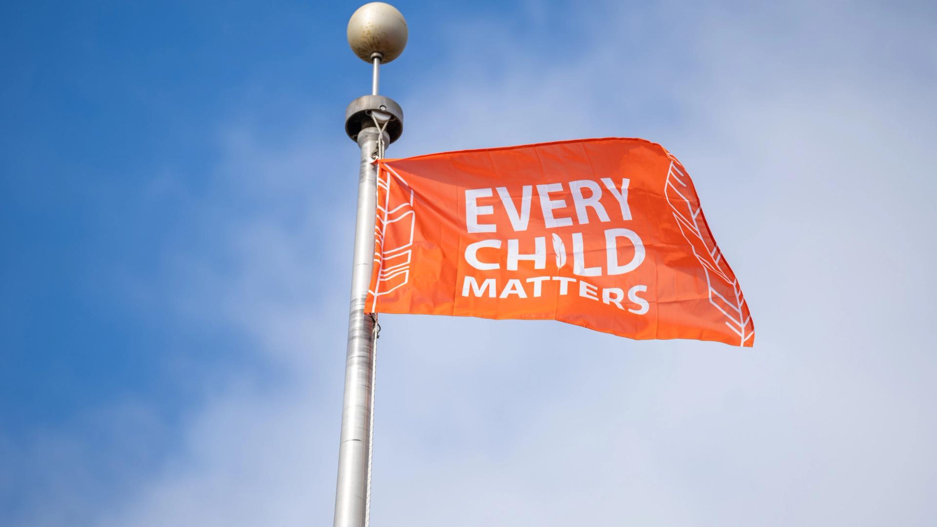 An orange flag reading “Every Child Matters” flies from a metal pole against a blue sky.