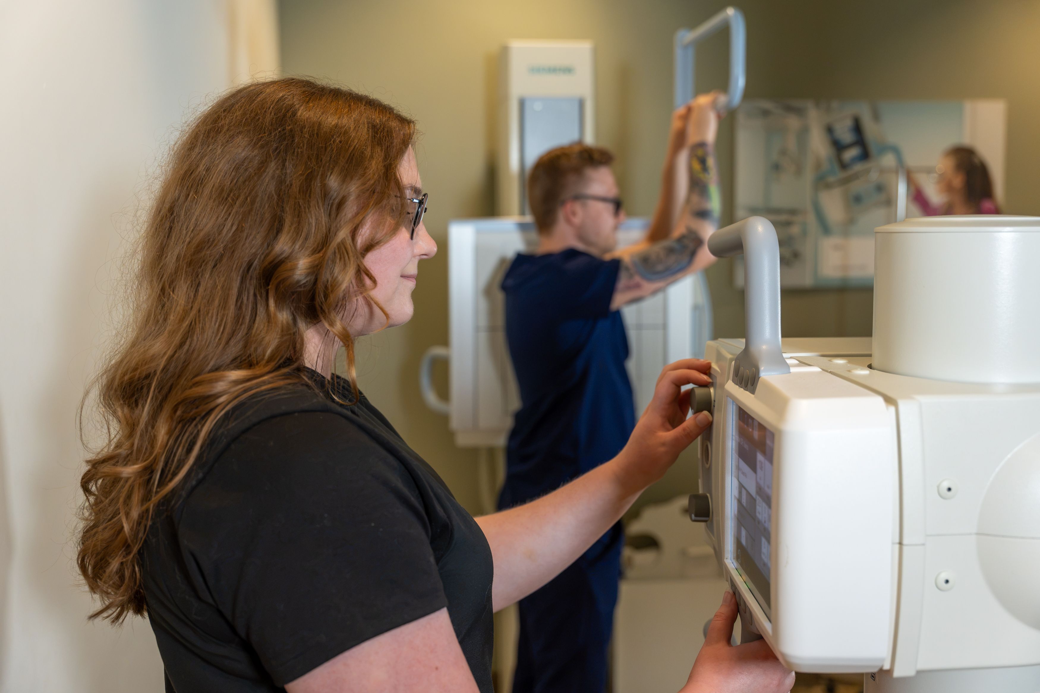 A woman adjusts the controls on medical imaging equipment while a man in navy scrubs handles a nearby machine.