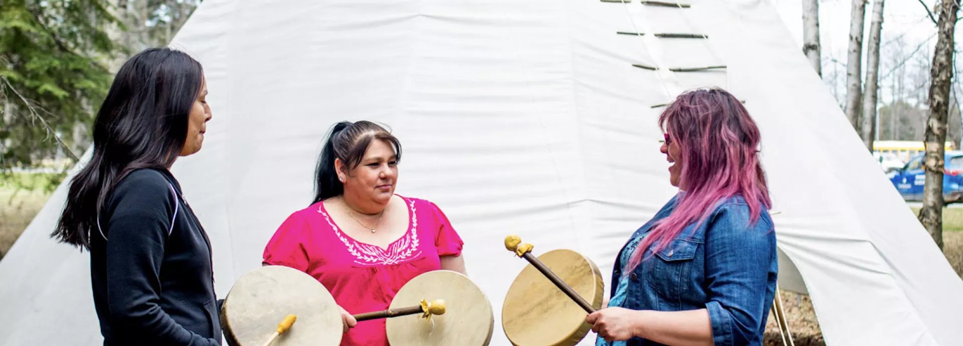 Three women standing in front of a large white tipi holding hand drums and talking together