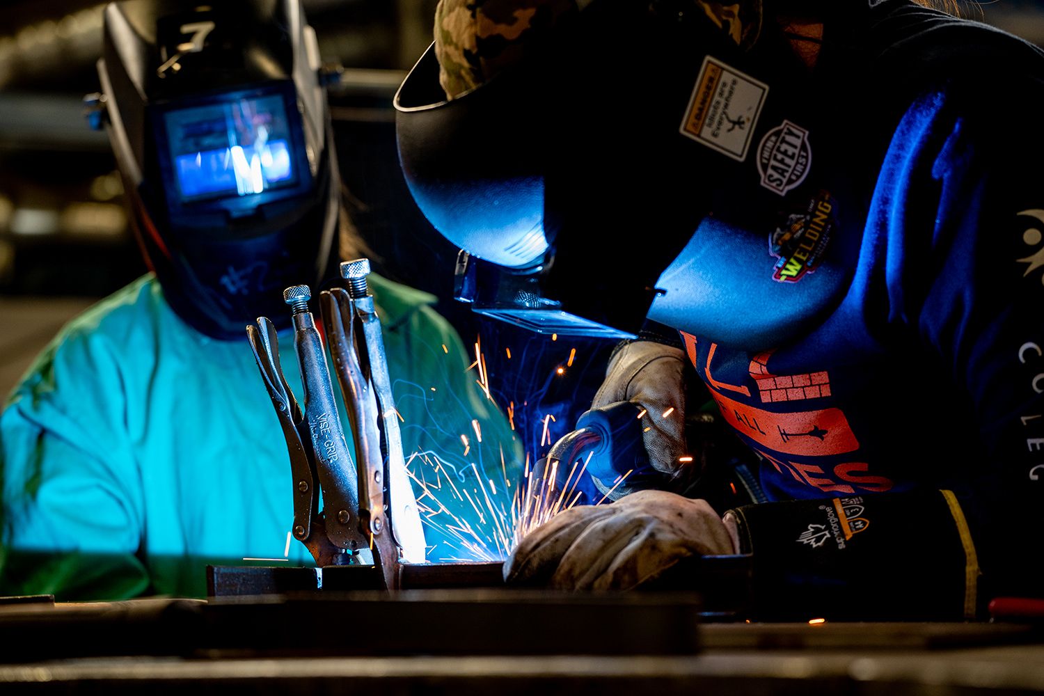 Two people wearing welding helmets and protective gloves work at a metal bench as bright sparks fly.