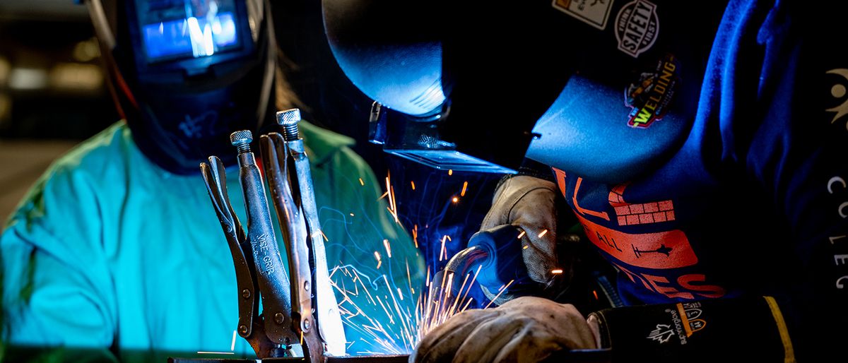 Two people wearing welding helmets and protective gloves work at a metal bench as bright sparks fly.