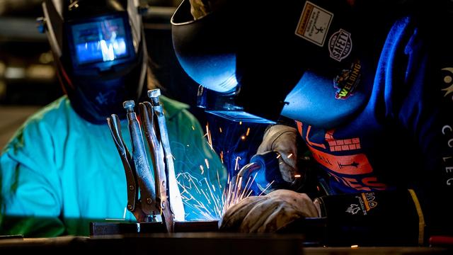 Two people wearing welding helmets and protective gloves work at a metal bench as bright sparks fly.