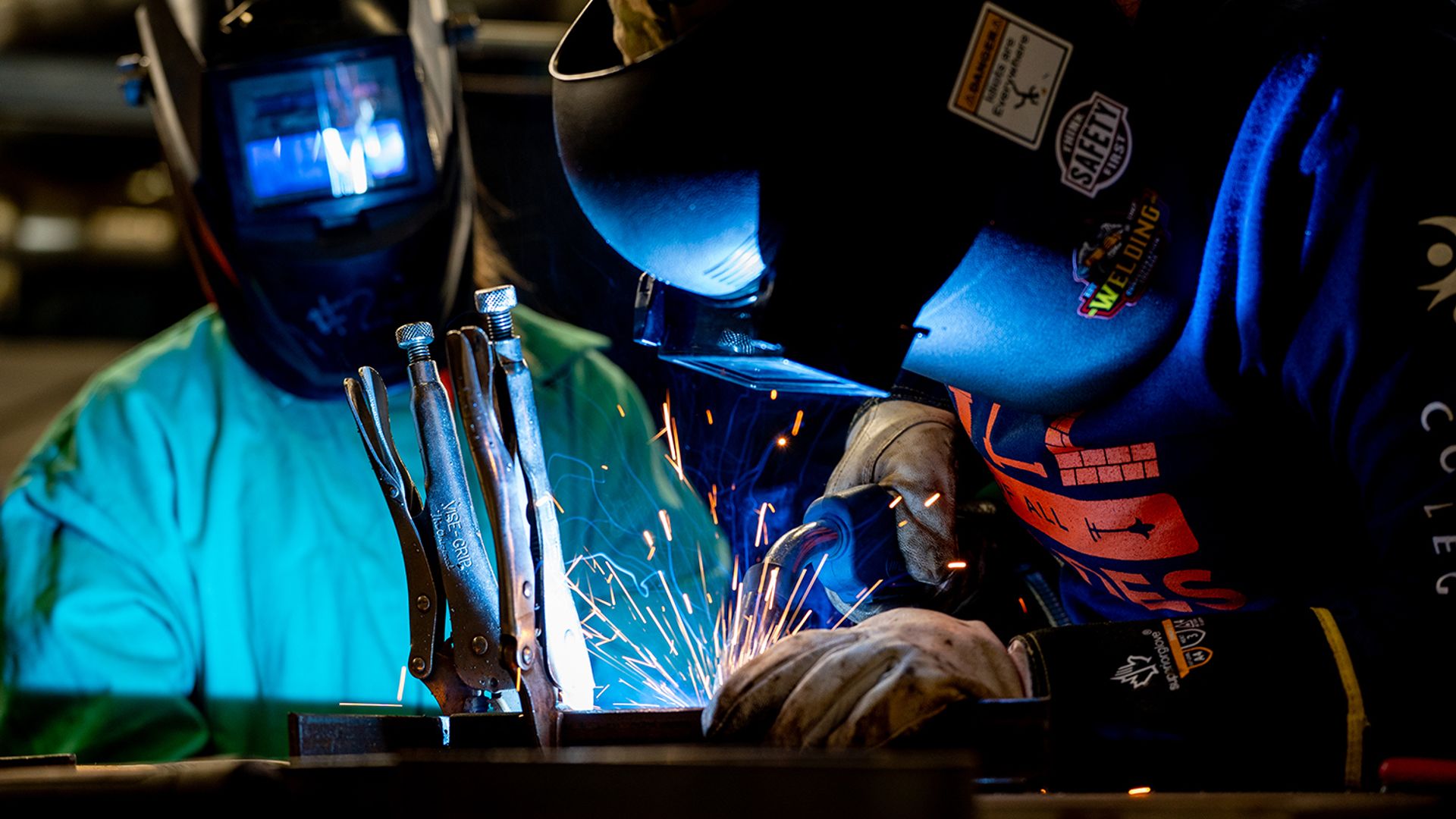 Two people wearing welding helmets and protective gloves work at a metal bench as bright sparks fly.
