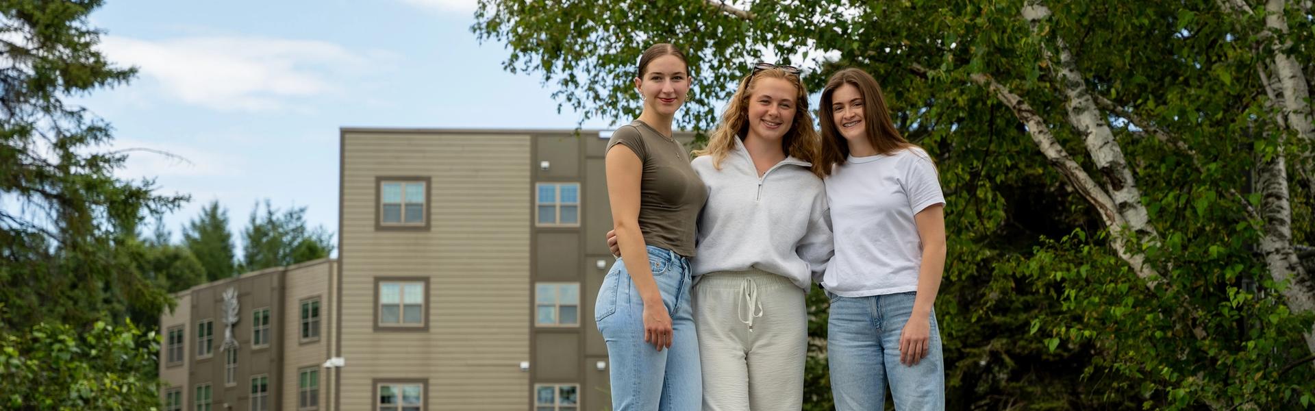 Three students stand together on a paved walkway outside a residence building on campus, posing casually with trees and grass around them