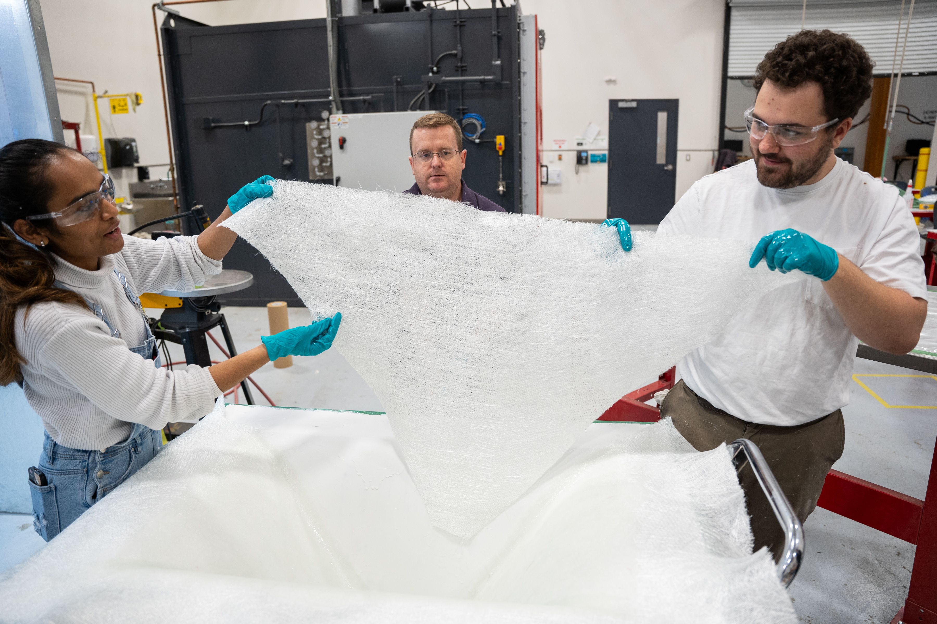 Three people in safety glasses and blue gloves hold a large white fibrous sheet in a workshop
