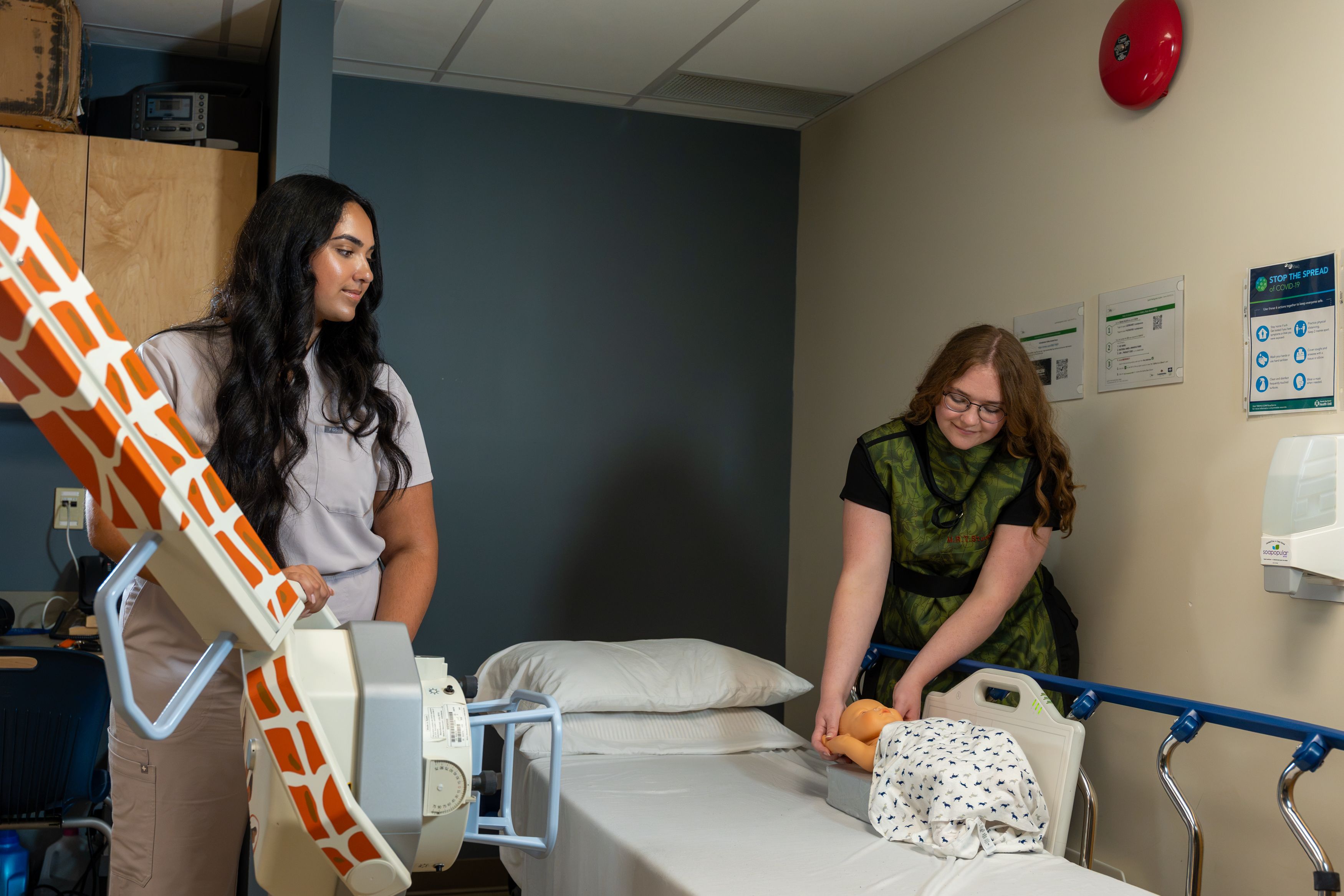 Two people in a clinical training room beside a bed, with one operating a large imaging machine and the other handling a mannequin.