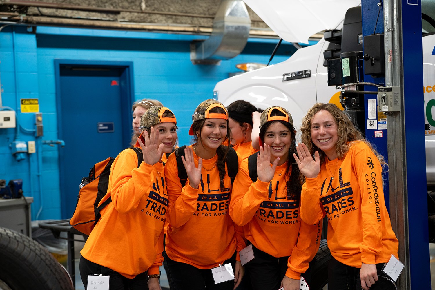 Four people in orange trades shirts and backwards caps wave in an automotive workshop beside a vehicle and equipment.