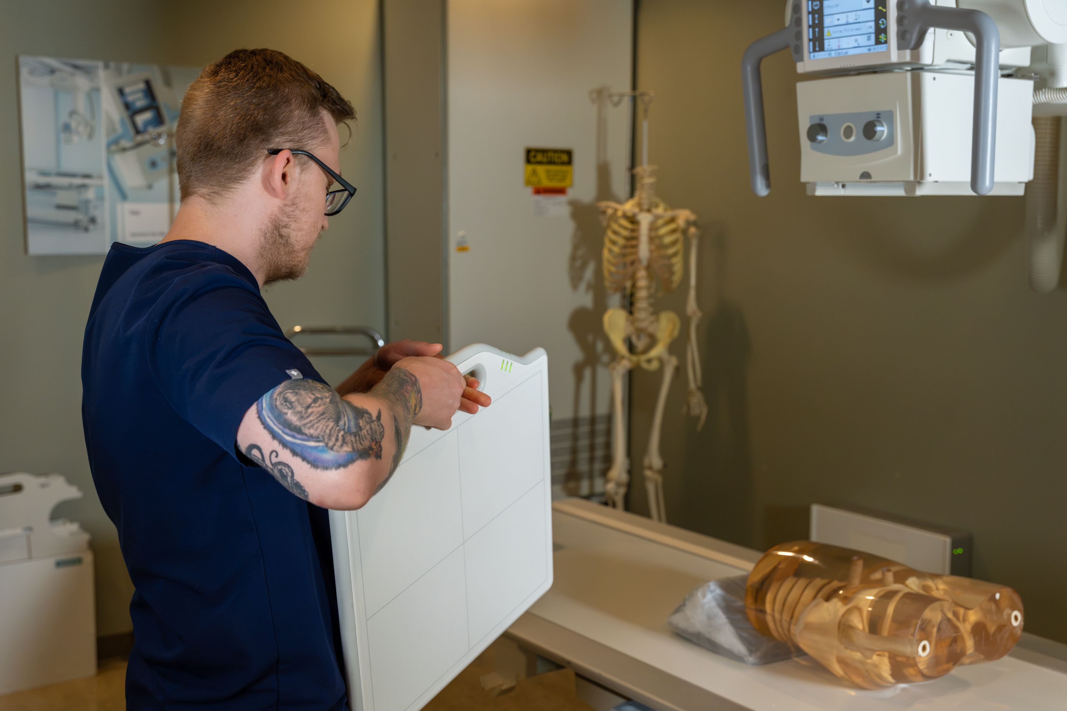 A person in navy scrubs operates a portable X-ray detector beside an X-ray machine and skeleton model.