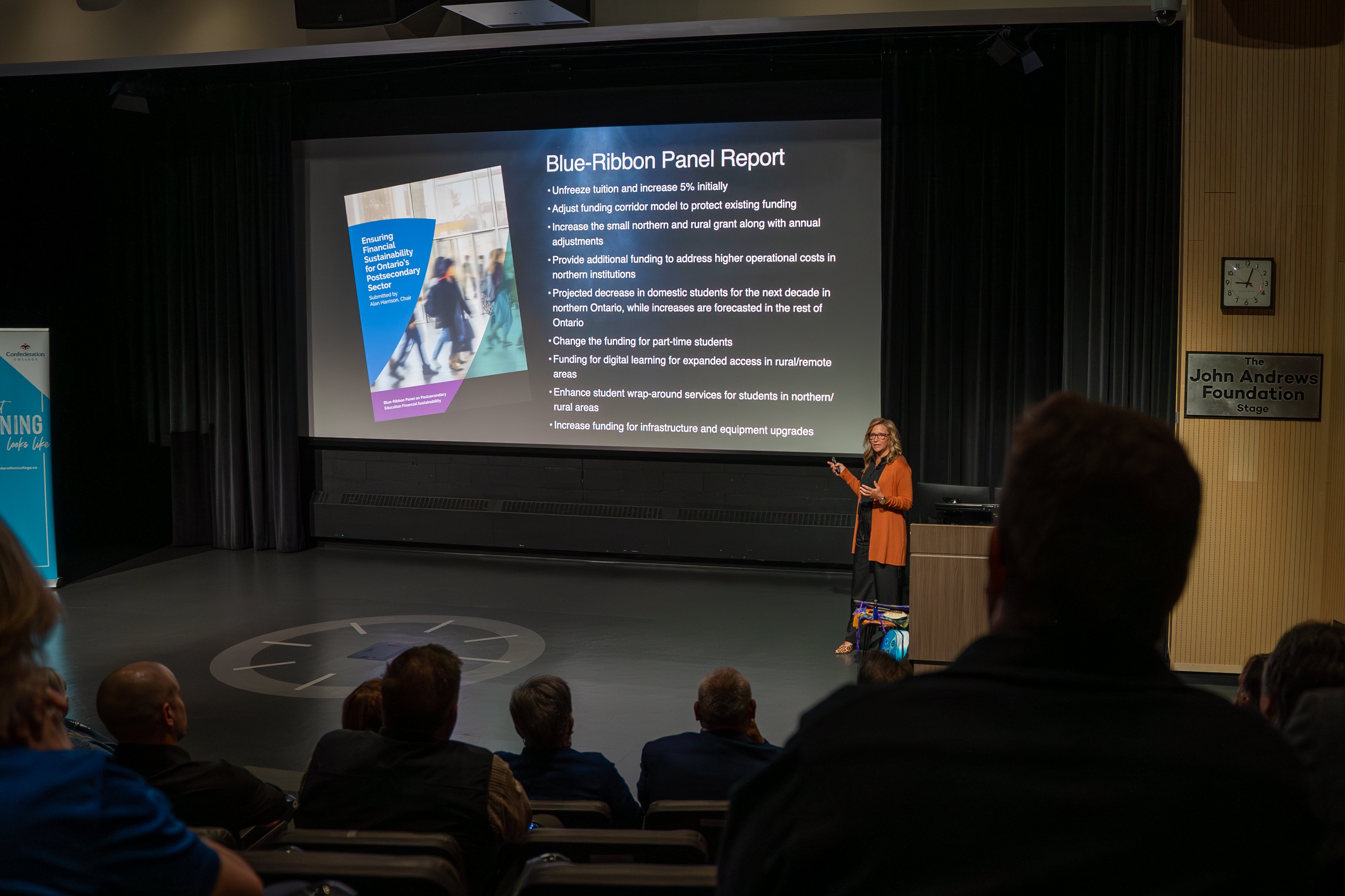 A speaker in an orange cardigan presents on stage at Confederation College, with a presentation titled 'Blue-Ribbon Panel Report' displayed on a large screen in a lecture hall.