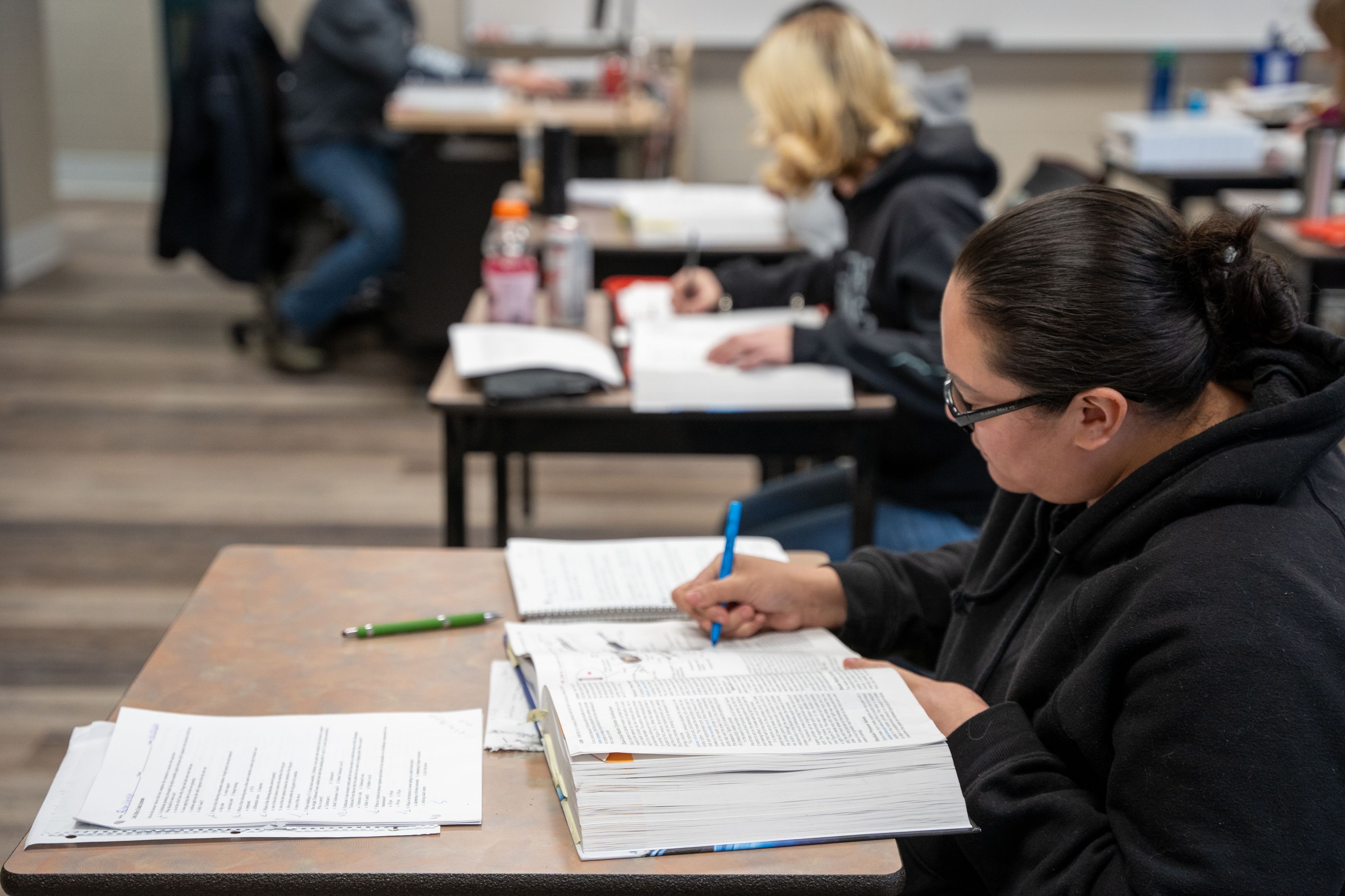 Several students sit at desks in a classroom, writing in notebooks and textbooks during a lesson