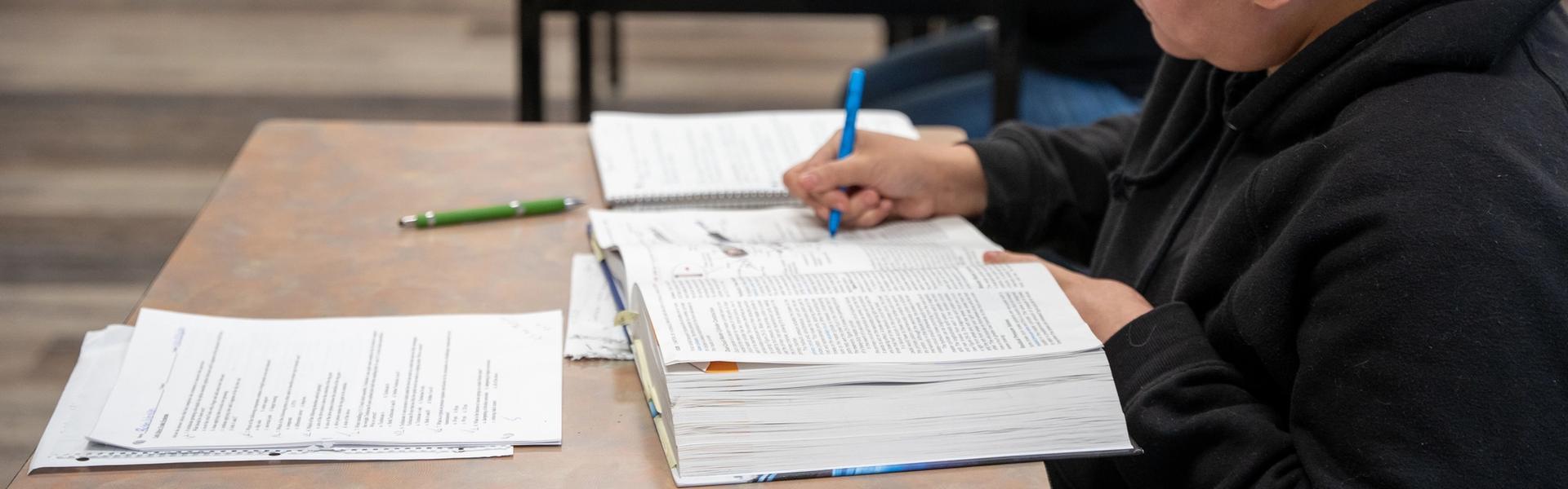 Several students sit at desks in a classroom, writing in notebooks and textbooks during a lesson