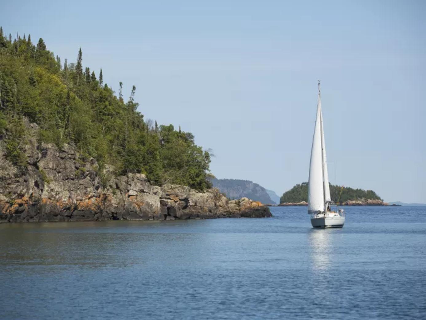 Sailboat gliding near rocky, tree-covered shoreline on a calm lake